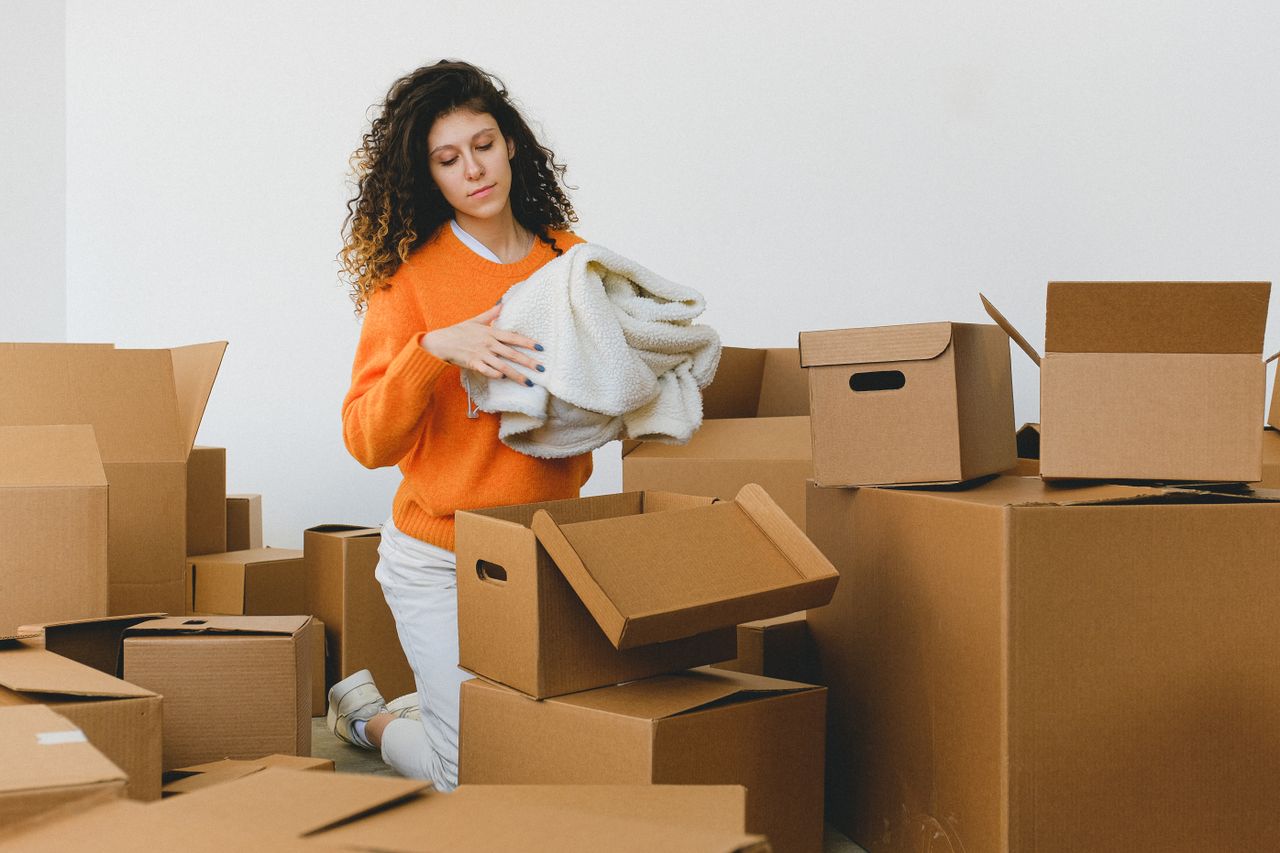 Services-01 Concentrated young lady with curly hair standing on knees on floor and packing clothes into carton boxes before moving into new flat